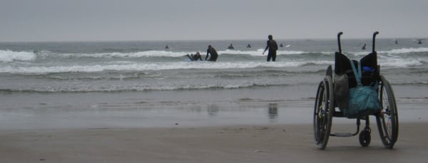 photo of beach and surfers with wheelchair in foreground