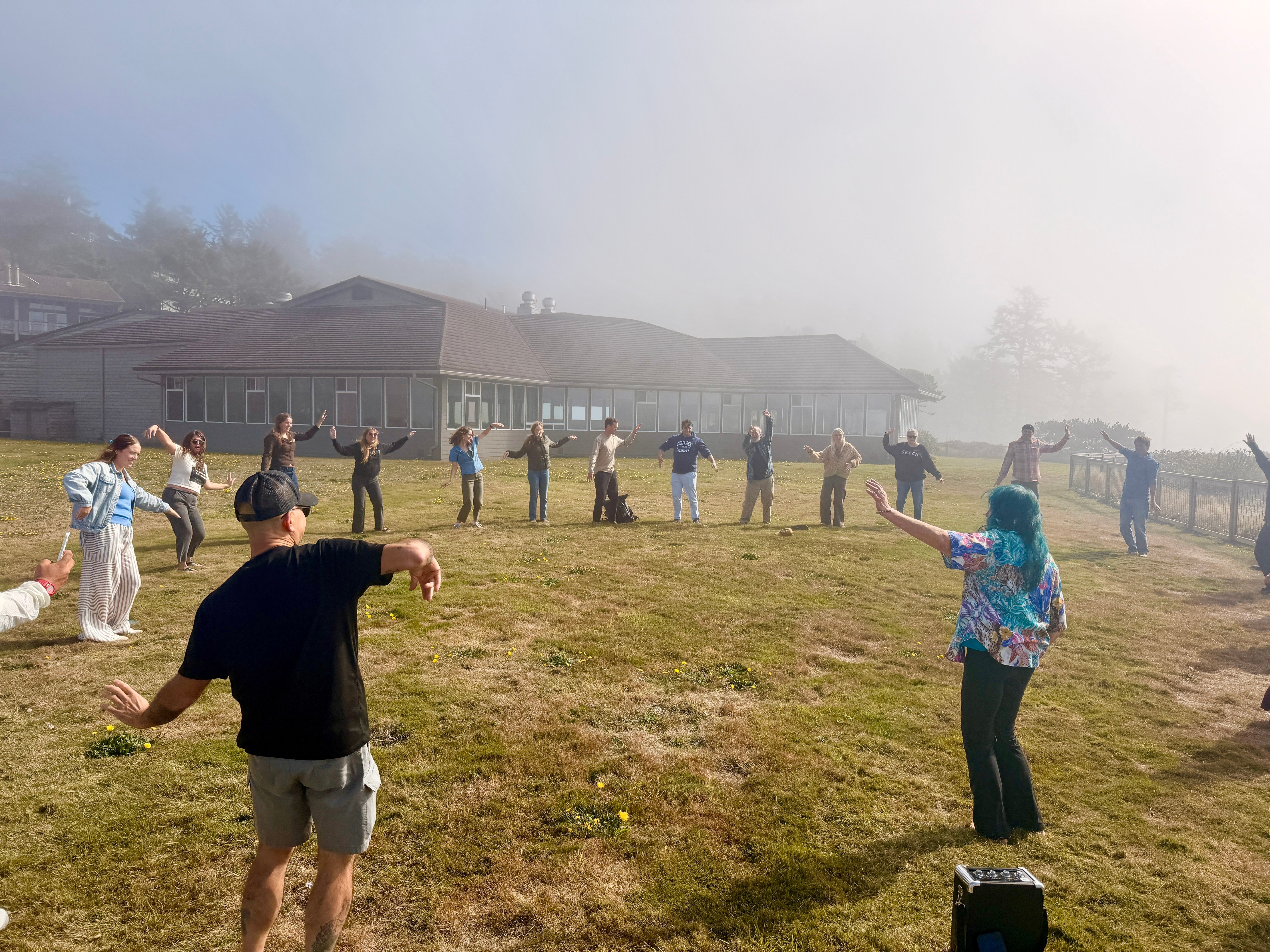 Volunteers gather in a circle outside on a misty lawn, practicing dance moves