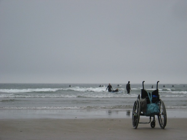 An empty wheelchair on a beach awaits while surfers ride the waves further off the beach